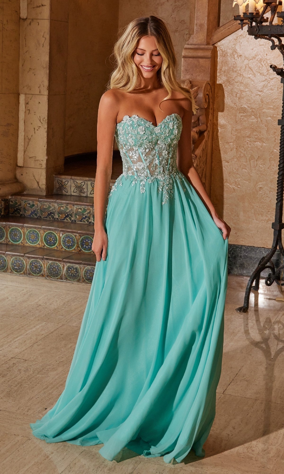 A woman stands indoors wearing the Nox Anabel C1617 Strapless Sweetheart Prom Dress in sage green, featuring a sheer embroidered bodice and a flowing, floor-length skirt.